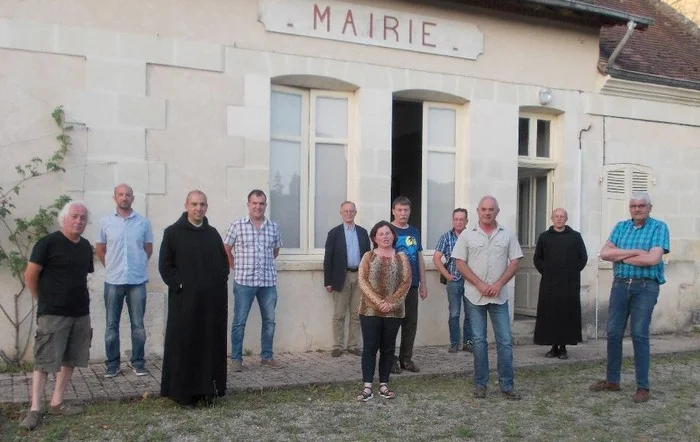 Photographie de groupe posant devant le bâtiment d'une mairie de village, réunissant une douzaine d'élus locaux en tenue civile et deux moines bénédictins vêtus de leur habit noir.
