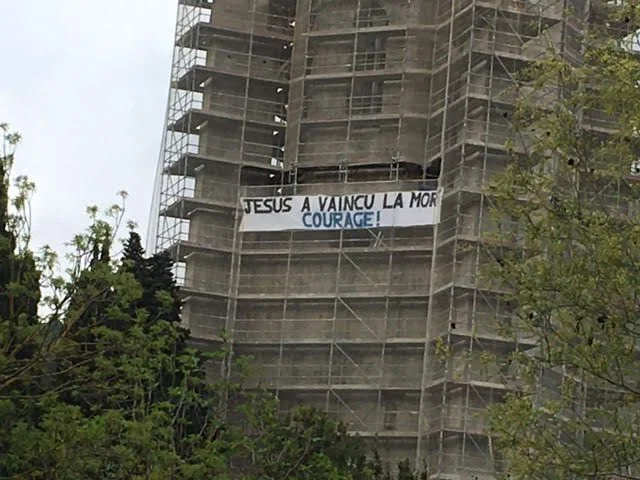 Photographie en gros plan du clocher de l'abbaye de Lagrasse entouré d'échafaudages métalliques pour travaux. Une grande banderole blanche accrochée aux structures porte le message « JESUS A VAINCU LA MOR » avec l'inscription « COURAGE ! » peinte en bleu juste en dessous.