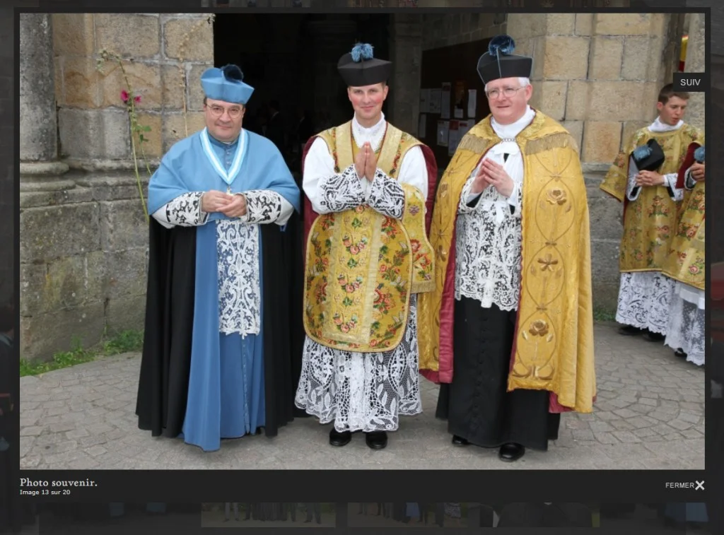 Trois ecclésiastiques membres de l&rsquo;Institut du Christ Roi Souverain Prêtre posent en vêtements liturgiques traditionnels devant un bâtiment en pierre. À gauche, Gilles Wach porte une mozette bleue avec une cape noire sur sa soutane. Au centre, Alban Denis en chasuble dorée brodée et aube en dentelle joint les mains. À droite, un prêtre est vêtu d&rsquo;une chape dorée sur un surplis en dentelle. Les trois hommes portent des barrettes cléricales sur la tête. Une légende en bas à gauche indique « Photo souvenir ».