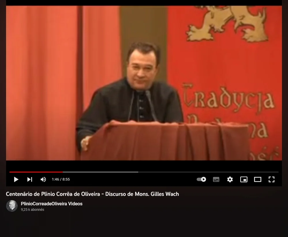 Mgr Gilles Wach prononçant un discours derrière un pupitre lors du centenaire de Plinio Corrêa de Oliveira. Il porte une soutane noire, tandis qu&rsquo;en arrière-plan un grand drapé rouge arbore un lion héraldique et l&rsquo;inscription « Tradycja ». En bas de l&rsquo;écran, les commandes du lecteur vidéo YouTube indiquent le titre de la vidéo et la chaîne « PlinioCorreadeOliveira Videos ».