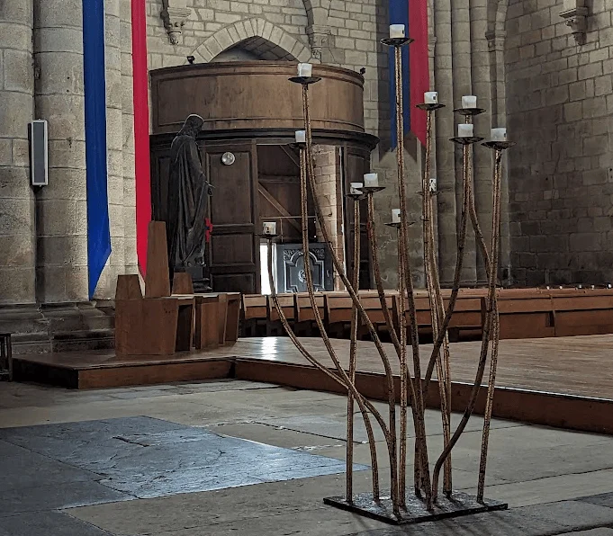 Grand candélabre métallique contemporain aux tiges ondulées supportant des bougies, placé au premier plan dans la collégiale de Saint-Yrieix-la-Perche, avec des sièges d&rsquo;officiants et une statue en arrière-plan.