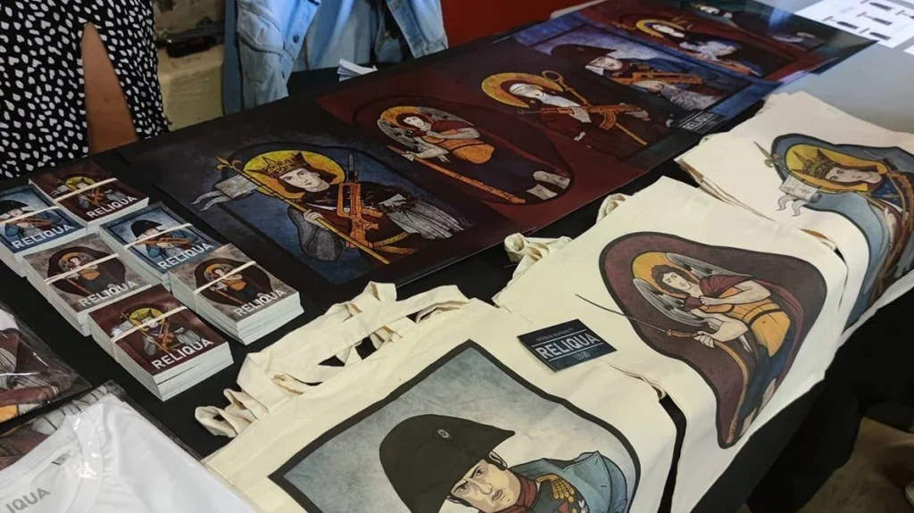 Stand d&rsquo;articles de la marque Reliqua à l&rsquo;université d&rsquo;été d&rsquo;Academia Christiana présentant des affiches, paquets de cartes, t-shirts et tote bags aux visuels détournés de figures historiques et religieuses armées.
