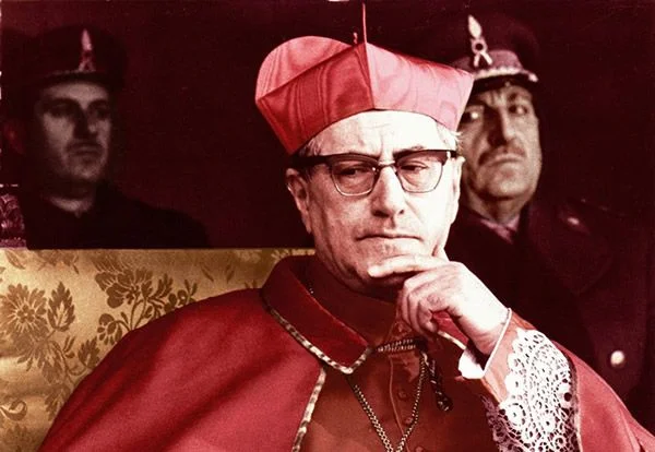 Le cardinal Giuseppe Siri, lunettes sur le nez et barrette rouge sur la tête, assis la main sous le menton devant deux hommes en uniforme en arrière-plan.