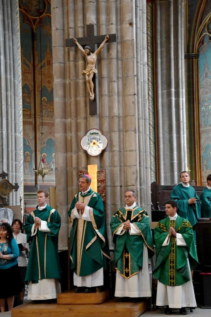 Célébration liturgique dans une église, montrant des clercs en chasubles vertes debout près d'un pilier en pierre surmonté d'un crucifix.