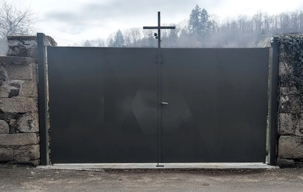 Grand portail métallique sombre plein, surmonté d&rsquo;une croix noire en son centre, encadré par deux piliers en murs de pierre avec une forêt en arrière-plan sous un ciel couvert.